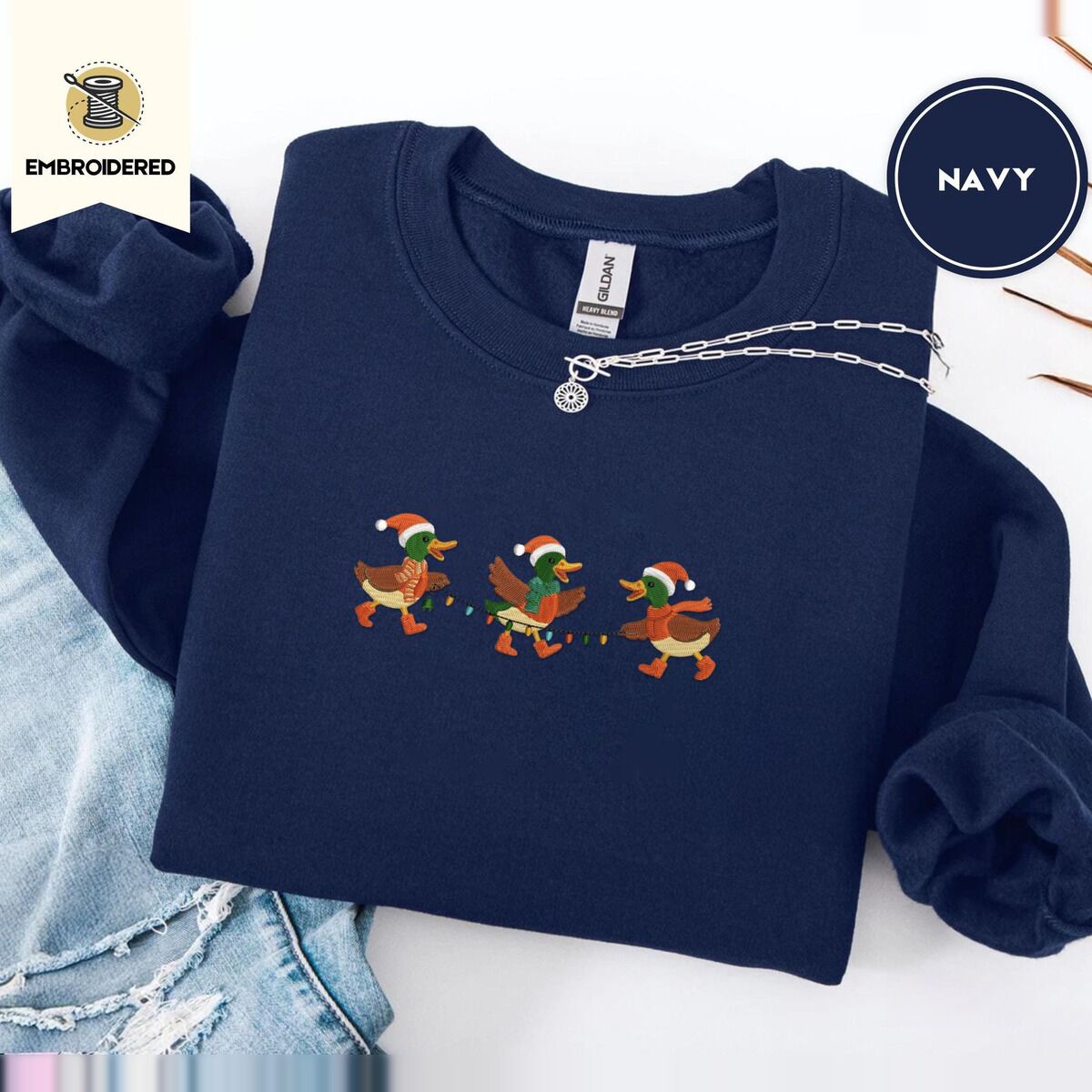 Mallard Duck Christmas Sweatshirt – Navy Navy crewneck sweatshirt folded with ripped jeans and a silver chain necklace, embroidered with three mallard ducks in Santa hats, knit scarves and orange boots carrying a string of Christmas lights