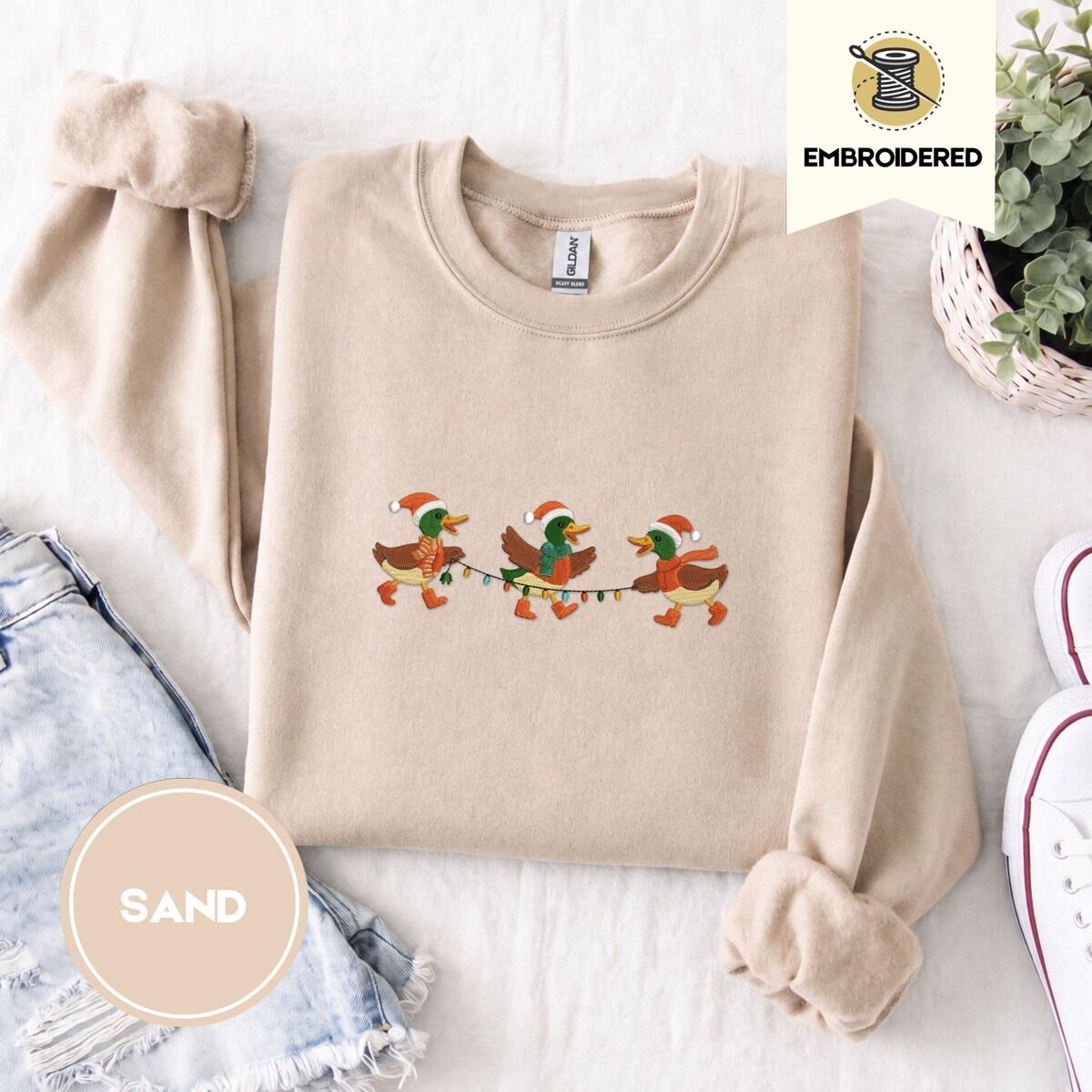 Mallard Duck Christmas Sweatshirt – Sand Sand crewneck sweatshirt folded with denim shorts, white sneakers and a potted plant, embroidered with three mallard ducks in Santa hats, knit scarves and orange boots carrying a string of Christmas lights