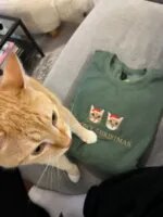 Green sweatshirt with two embroidered cat faces in Santa hats and Meowy Christmas text, next to an orange cat
