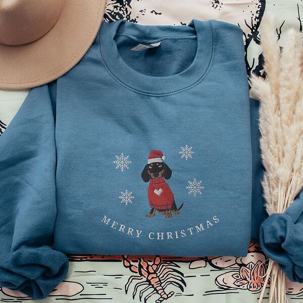 Indigo Blue crewneck sweatshirt laid flat with a felt hat and pampas grass, embroidered with a black and tan dachshund in a Santa hat and red heart sweater among snowflakes above the words Merry Christmas