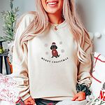 Woman sitting in a holiday-decorated room in a Sand crewneck sweatshirt, embroidered with a black and tan dachshund in a Santa hat and red heart sweater among snowflakes above the words Merry Christmas