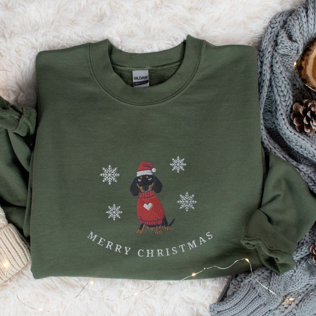Merry Christmas Dachshund Sweatshirt – Military Green Military Green crewneck sweatshirt laid flat on a fur rug beside a knit blanket and pinecones, embroidered with a black and tan dachshund in a Santa hat and red heart sweater among snowflakes above the words Merry Christmas