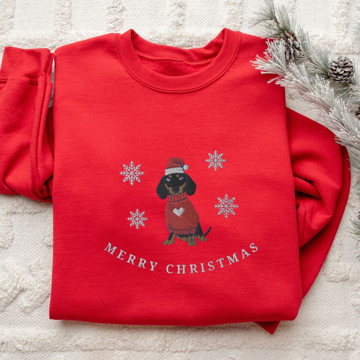 Merry Christmas Dachshund Sweatshirt - Red Red crewneck sweatshirt laid flat on a white knit blanket beside pine branches, embroidered with a black and tan dachshund in a Santa hat and red heart sweater among snowflakes above the words Merry Christmas