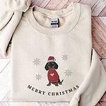 Sand crewneck sweatshirt laid flat on a wood floor, embroidered with a black and tan dachshund in a Santa hat and red heart sweater among snowflakes above the words Merry Christmas