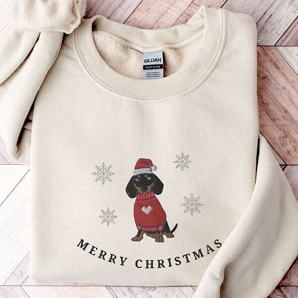 Sand crewneck sweatshirt laid flat on a wood floor, embroidered with a black and tan dachshund in a Santa hat and red heart sweater among snowflakes above the words Merry Christmas
