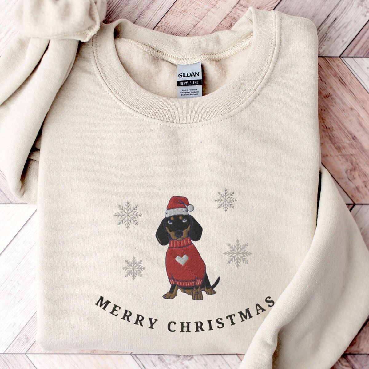 Merry Christmas Dachshund Sweatshirt – Sand Sand crewneck sweatshirt laid flat on a wood floor, embroidered with a black and tan dachshund in a Santa hat and red heart sweater among snowflakes above the words Merry Christmas