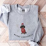 Sport Grey crewneck sweatshirt laid flat on a wood floor, embroidered with a black and tan dachshund in a Santa hat and red heart sweater among snowflakes above the words Merry Christmas