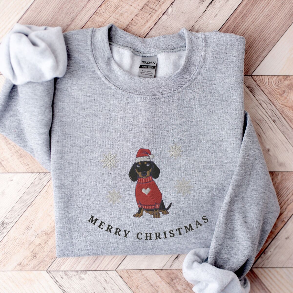 Merry Christmas Dachshund Sweatshirt - Sport Grey Sport Grey crewneck sweatshirt laid flat on a wood floor, embroidered with a black and tan dachshund in a Santa hat and red heart sweater among snowflakes above the words Merry Christmas