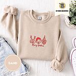 Sand crewneck sweatshirt folded with denim shorts, white sneakers and a potted plant, embroidered with three red line-art dog faces, a beagle in reindeer antlers, a retriever in a Santa hat and a pug in a party hat, above Merry Woofmas in script