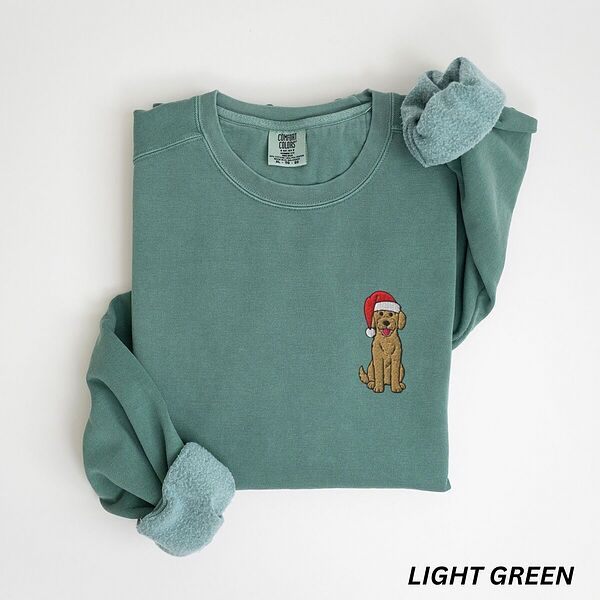 Mini goldendoodle Christmas sweatshirt in Light Green laid flat with rolled cuffs, embroidered with a small doodle in a red Santa hat on the left chest