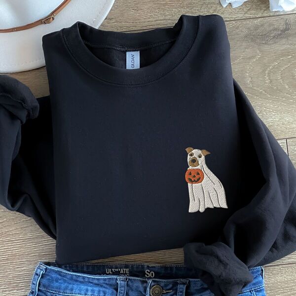 Black crewneck sweatshirt laid flat on a wood floor with jeans and a felt hat, embroidered with a small pit bull in a ghost sheet holding a jack-o'-lantern bucket on the left chest