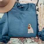 Indigo Blue crewneck sweatshirt laid flat on a patterned throw with a felt hat and pampas grass, embroidered with a small pit bull in a ghost sheet holding a jack-o'-lantern bucket on the left chest