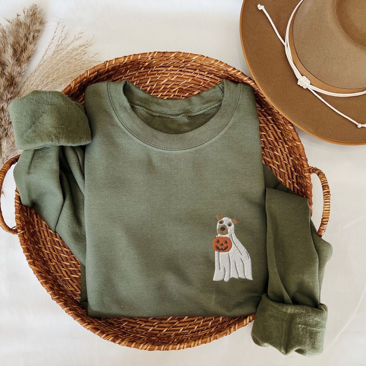 Mini Pitbull Ghost Sweatshirt – Military Green Military Green crewneck sweatshirt folded in a woven basket next to a felt hat, embroidered with a small pit bull in a ghost sheet holding a jack-o'-lantern bucket on the left chest