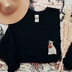 Black crewneck sweatshirt laid flat with a felt hat and pampas grass, embroidered with a small pit bull in a ghost sheet holding a jack-o'-lantern bucket on the left chest