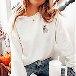Woman sitting by pumpkins in a white crewneck sweatshirt, holding a cup, embroidered with a small pit bull in a ghost sheet holding a jack-o'-lantern bucket on the left chest