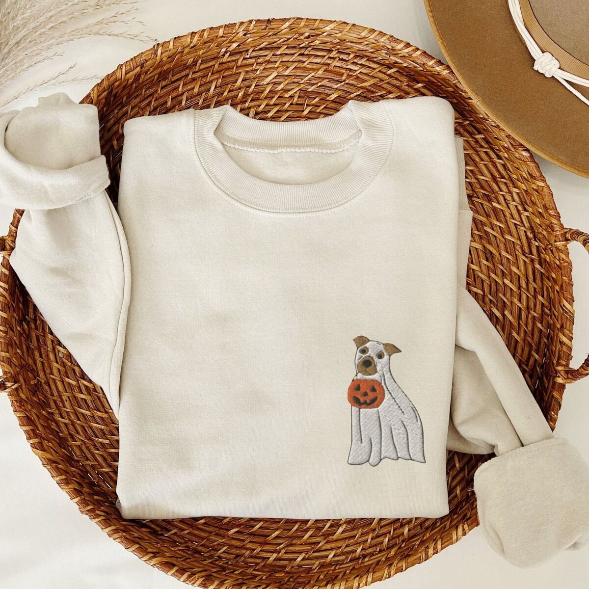 Mini Pitbull Ghost Sweatshirt – Sand Sand crewneck sweatshirt folded in a woven basket, embroidered with a small pit bull in a ghost sheet holding a jack-o'-lantern bucket on the left chest