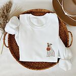 White crewneck sweatshirt folded in a woven basket with dried grass and a felt hat, embroidered with a small pit bull in a ghost sheet holding a jack-o'-lantern bucket on the left chest