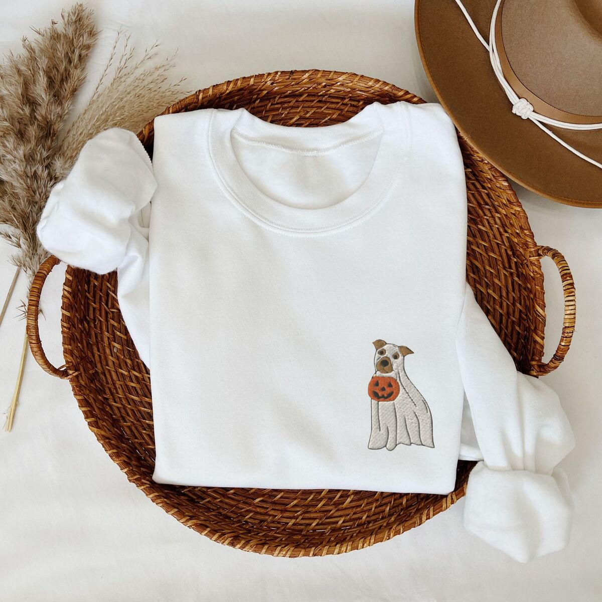 Mini Pitbull Ghost Sweatshirt - White White crewneck sweatshirt folded in a woven basket with dried grass and a felt hat, embroidered with a small pit bull in a ghost sheet holding a jack-o'-lantern bucket on the left chest