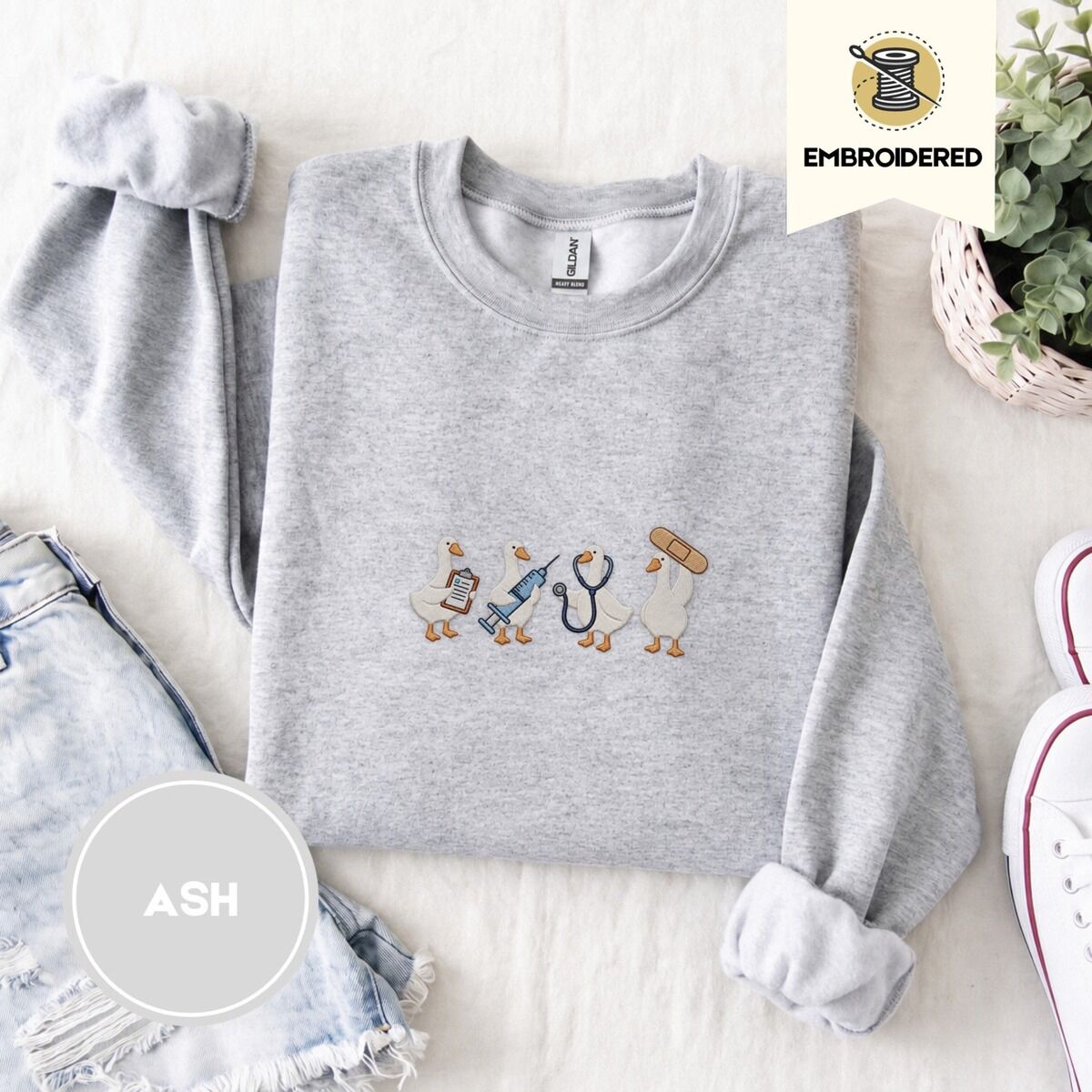Nurse Duck Sweatshirt – Ash Ash crewneck sweatshirt folded with denim shorts, white sneakers and a potted plant, embroidered with four white ducks holding a clipboard, a syringe, a stethoscope and a bandage