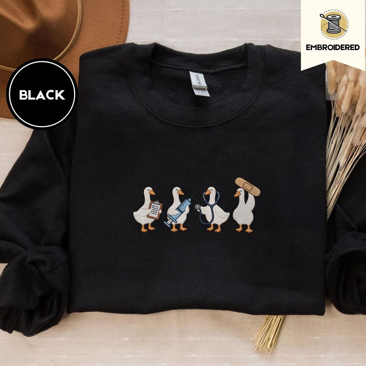 Nurse Duck Sweatshirt – Black Black crewneck sweatshirt with a brown felt hat and dried bunny tail grass, embroidered with four white ducks holding a clipboard, a syringe, a stethoscope and a bandage