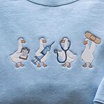 Close-up of four embroidered white ducks on a Light Blue sweatshirt: one with a clipboard, one with a blue syringe, one wearing a navy stethoscope and one lifting a tan bandage