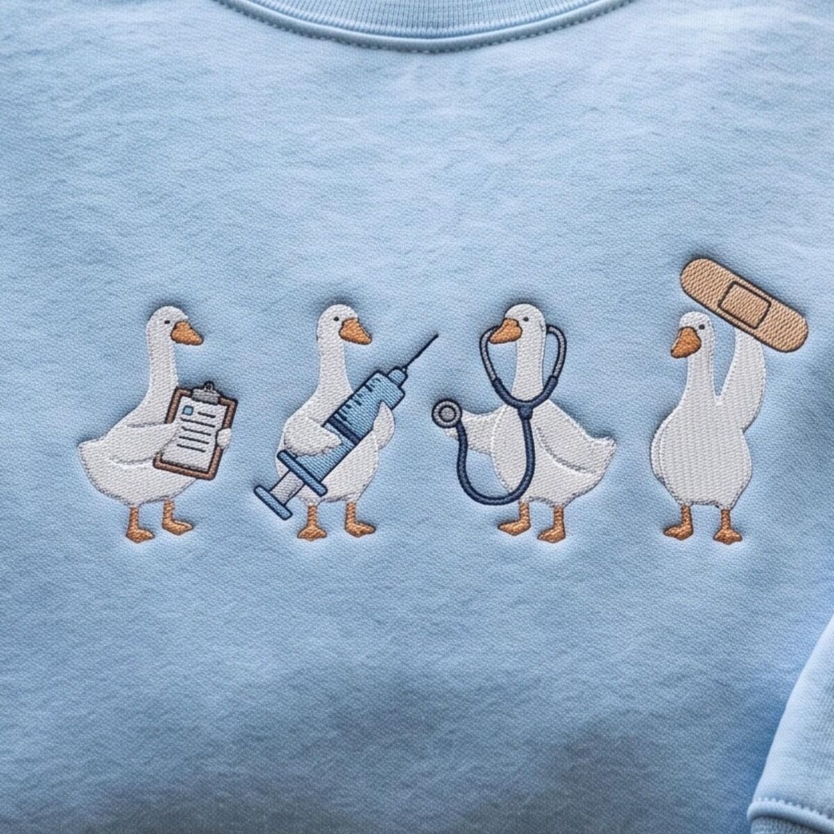 Nurse Duck Sweatshirt – Embroidery Close-Up Close-up of four embroidered white ducks on a Light Blue sweatshirt: one with a clipboard, one with a blue syringe, one wearing a navy stethoscope and one lifting a tan bandage