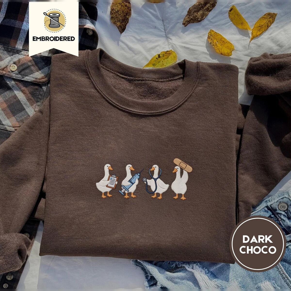 Nurse Duck Sweatshirt – Dark Chocolate Dark Chocolate crewneck sweatshirt on a plaid flannel shirt with yellow autumn leaves and denim shorts, embroidered with four white ducks holding a clipboard, a syringe, a stethoscope and a bandage