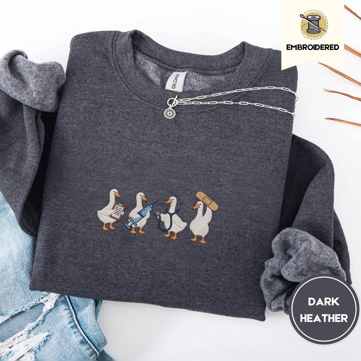 Nurse Duck Sweatshirt – Dark Heather Dark Heather crewneck sweatshirt folded with ripped jeans and a silver chain necklace, embroidered with four white ducks holding a clipboard, a syringe, a stethoscope and a bandage