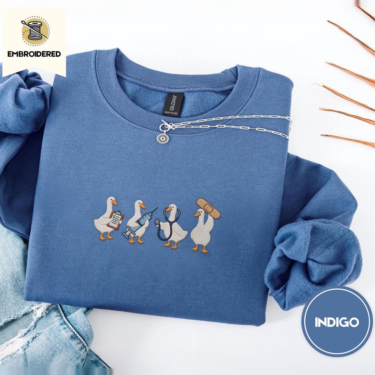 Nurse Duck Sweatshirt – Indigo Blue Indigo Blue crewneck sweatshirt folded with light jeans and dried twigs, embroidered with four white ducks holding a clipboard, a syringe, a stethoscope and a bandage