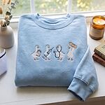 Light Blue crewneck sweatshirt folded on a white windowsill with dried bunny tail grass, a candle and old books, embroidered with four white ducks holding a clipboard, a syringe, a stethoscope and a bandage