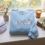 Light Blue crewneck sweatshirt on a windowsill beside a nurse cap, a candle, a stethoscope and a nursing journal, embroidered with four medical ducks across the chest