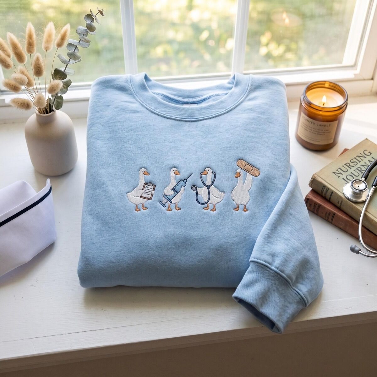 Nurse Duck Sweatshirt – Light Blue with a Nurse Cap Light Blue crewneck sweatshirt on a windowsill beside a nurse cap, a candle, a stethoscope and a nursing journal, embroidered with four medical ducks across the chest