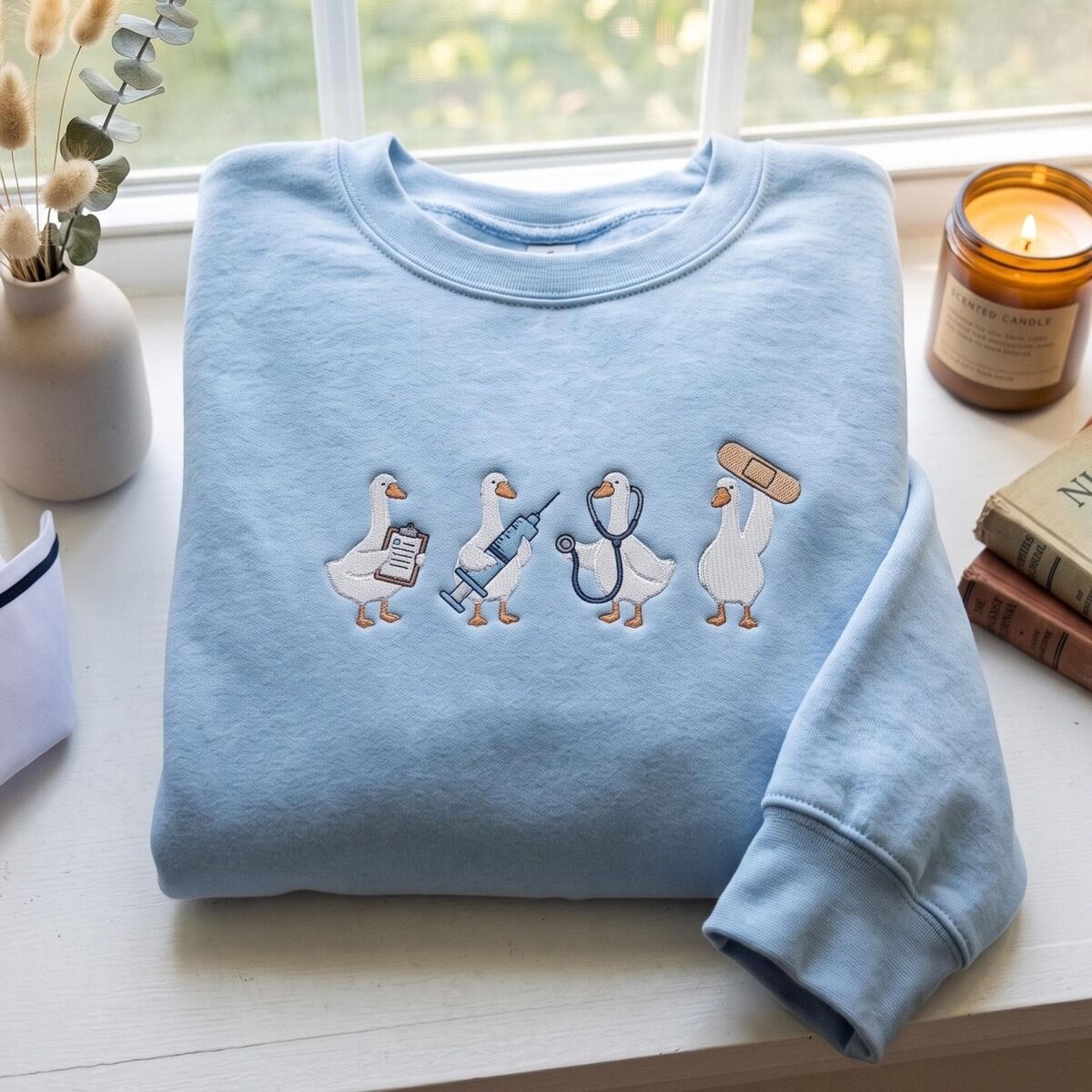 Nurse Duck Sweatshirt – Light Blue Light Blue crewneck sweatshirt folded on a white windowsill with dried bunny tail grass, a candle and old books, embroidered with four white ducks holding a clipboard, a syringe, a stethoscope and a bandage