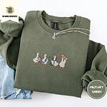 Military Green crewneck sweatshirt folded with light jeans and a silver chain necklace, embroidered with four white ducks holding a clipboard, a syringe, a stethoscope and a bandage
