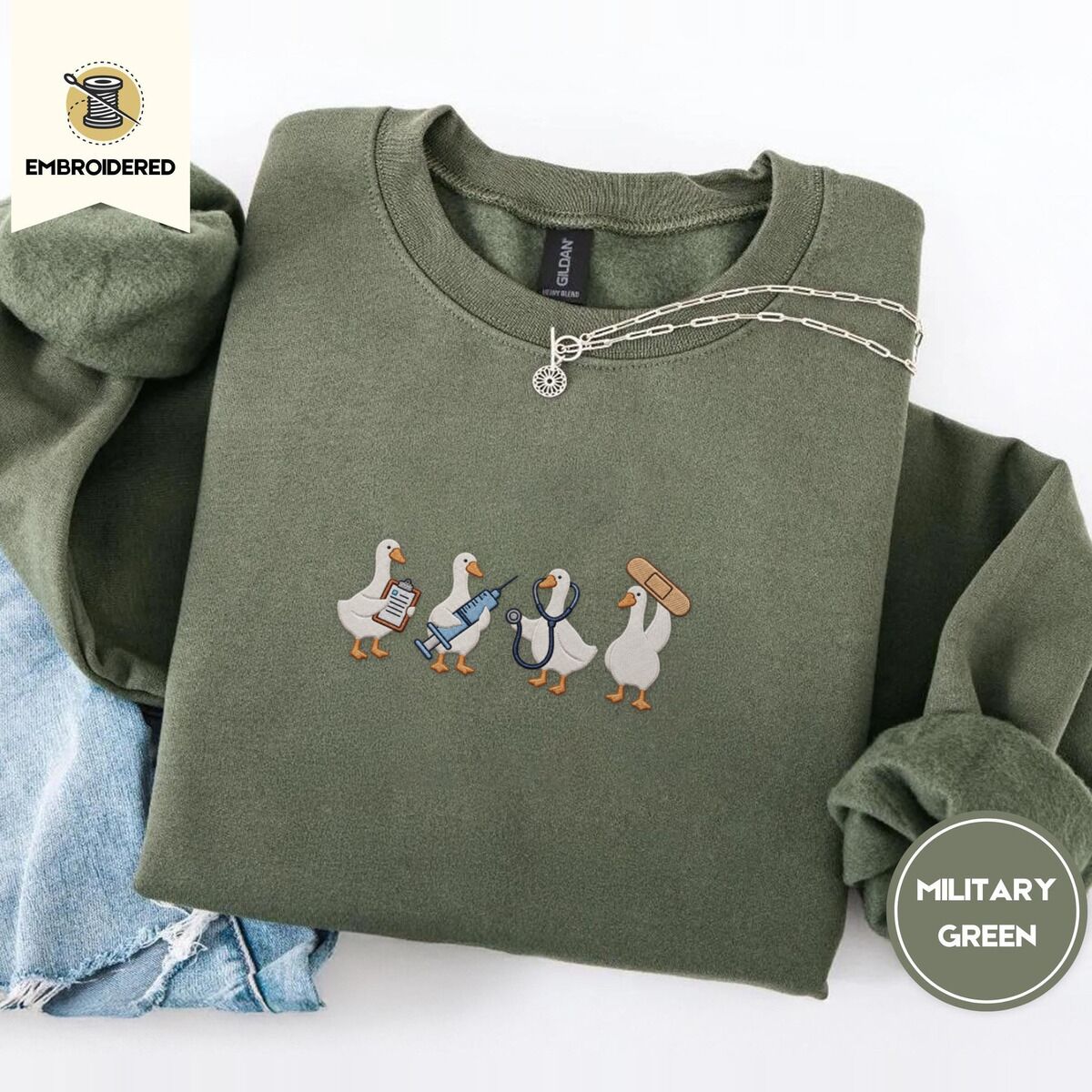 Nurse Duck Sweatshirt – Military Green Military Green crewneck sweatshirt folded with light jeans and a silver chain necklace, embroidered with four white ducks holding a clipboard, a syringe, a stethoscope and a bandage