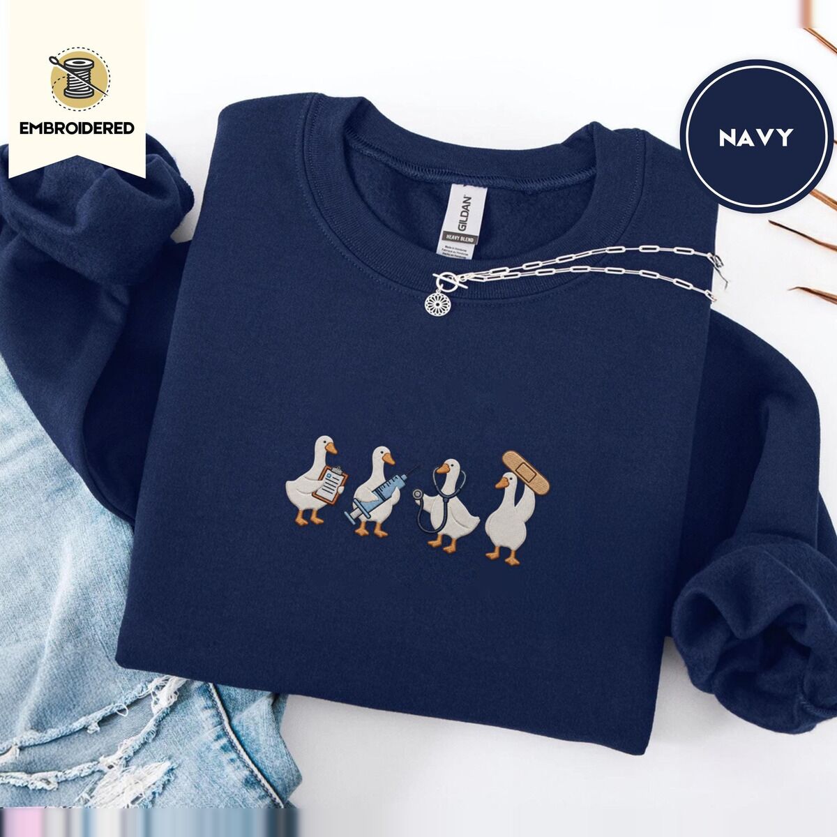 Nurse Duck Sweatshirt – Navy Navy crewneck sweatshirt folded with ripped jeans and a silver chain necklace, embroidered with four white ducks holding a clipboard, a syringe, a stethoscope and a bandage