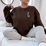 Woman sitting on a bed in a Dark Chocolate crewneck sweatshirt embroidered with a shaggy Old English sheepdog sitting in profile with a white front, a dark back and a red tongue on the left chest