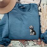 Indigo Blue crewneck sweatshirt laid flat with a tan hat and dried pampas grass, embroidered with a shaggy Old English sheepdog sitting in profile with a white front, a dark back and a red tongue on the left chest
