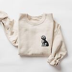 Sand crewneck sweatshirt laid flat on a white background, embroidered with a shaggy Old English sheepdog sitting in profile with a white front, a dark back and a red tongue on the left chest