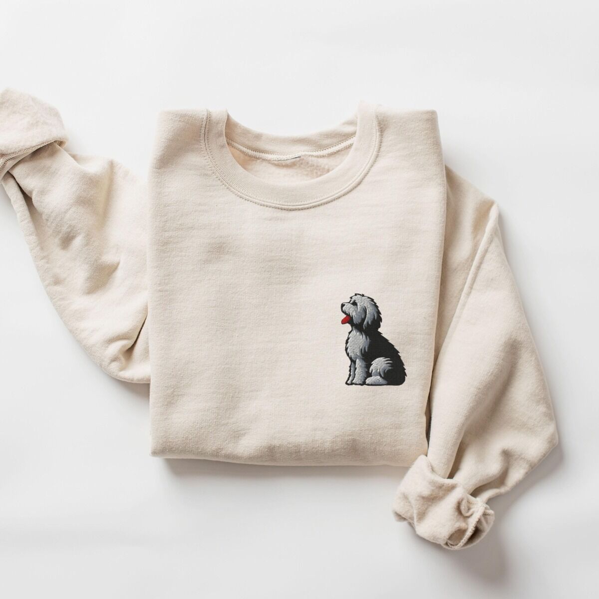 Old English Sheepdog Sweatshirt – Sand Sand crewneck sweatshirt laid flat on a white background, embroidered with a shaggy Old English sheepdog sitting in profile with a white front, a dark back and a red tongue on the left chest