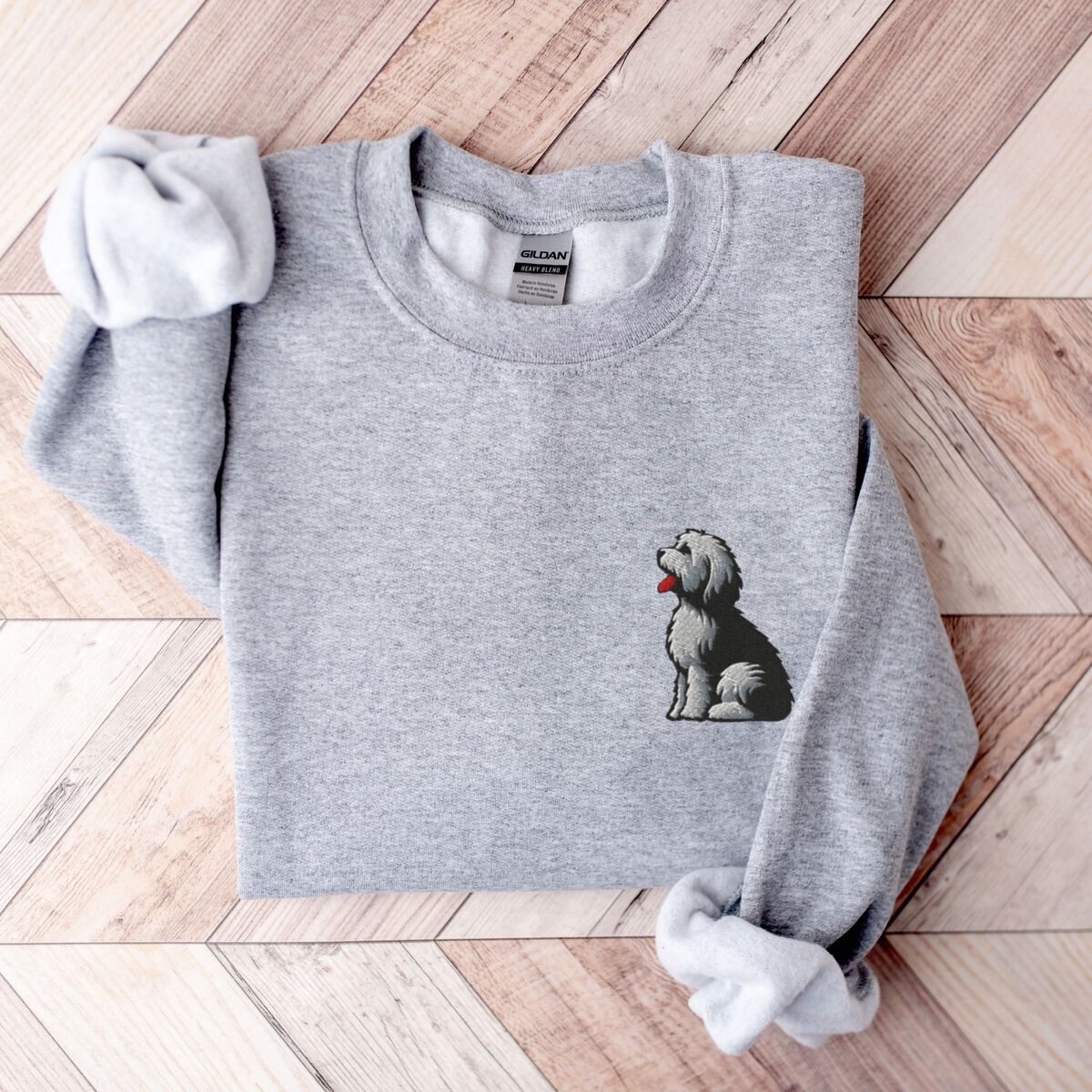 Old English Sheepdog Sweatshirt - Sport Grey Sport Grey crewneck sweatshirt laid flat on a herringbone wood floor, embroidered with a shaggy Old English sheepdog sitting in profile with a white front, a dark back and a red tongue on the left chest