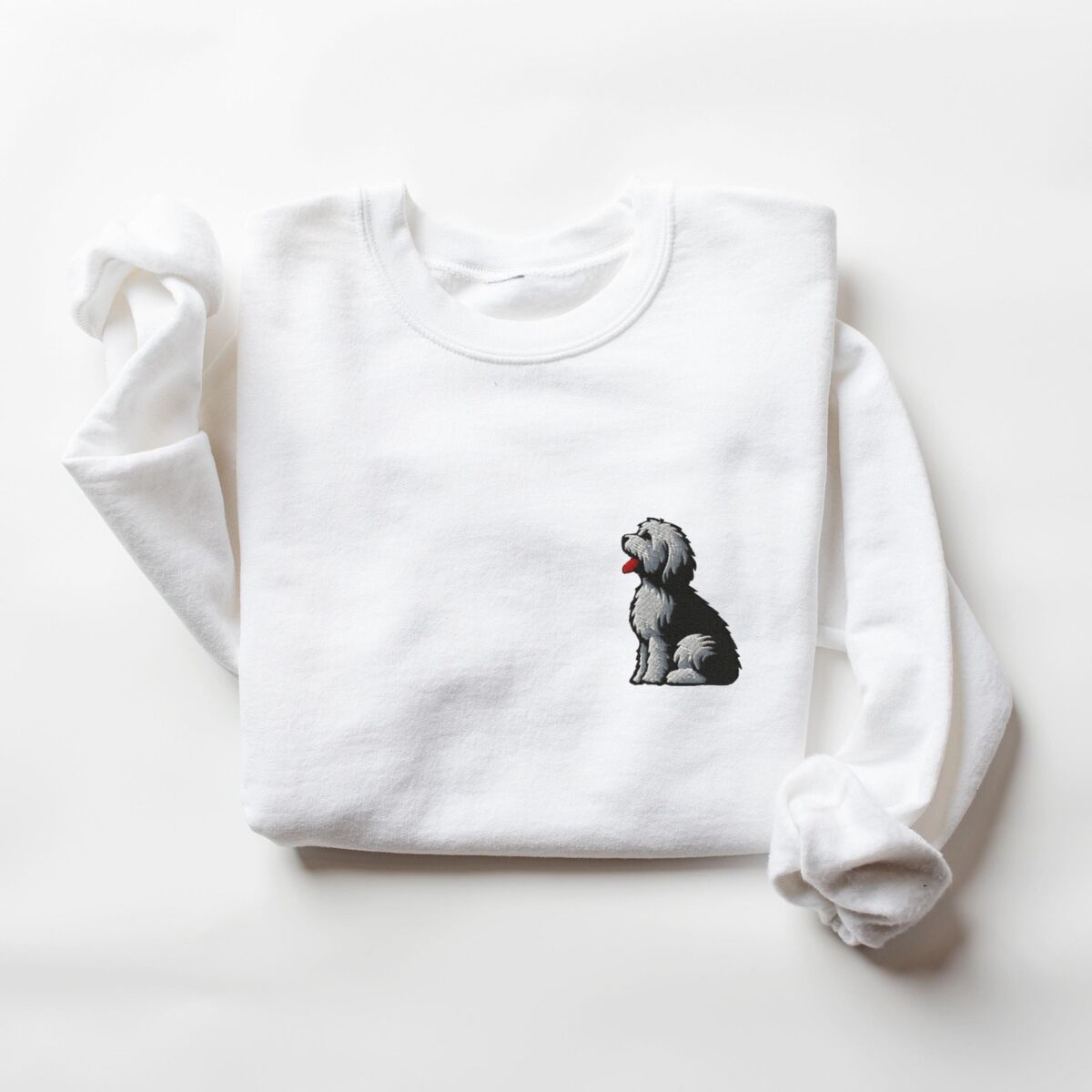 Old English Sheepdog Sweatshirt - White White crewneck sweatshirt laid flat on a white background, embroidered with a shaggy Old English sheepdog sitting in profile with a white front, a dark back and a red tongue on the left chest