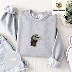 Ash crewneck sweatshirt folded with denim shorts, white sneakers and a potted plant, embroidered with a river otter head in brown and cream thread with long golden whiskers