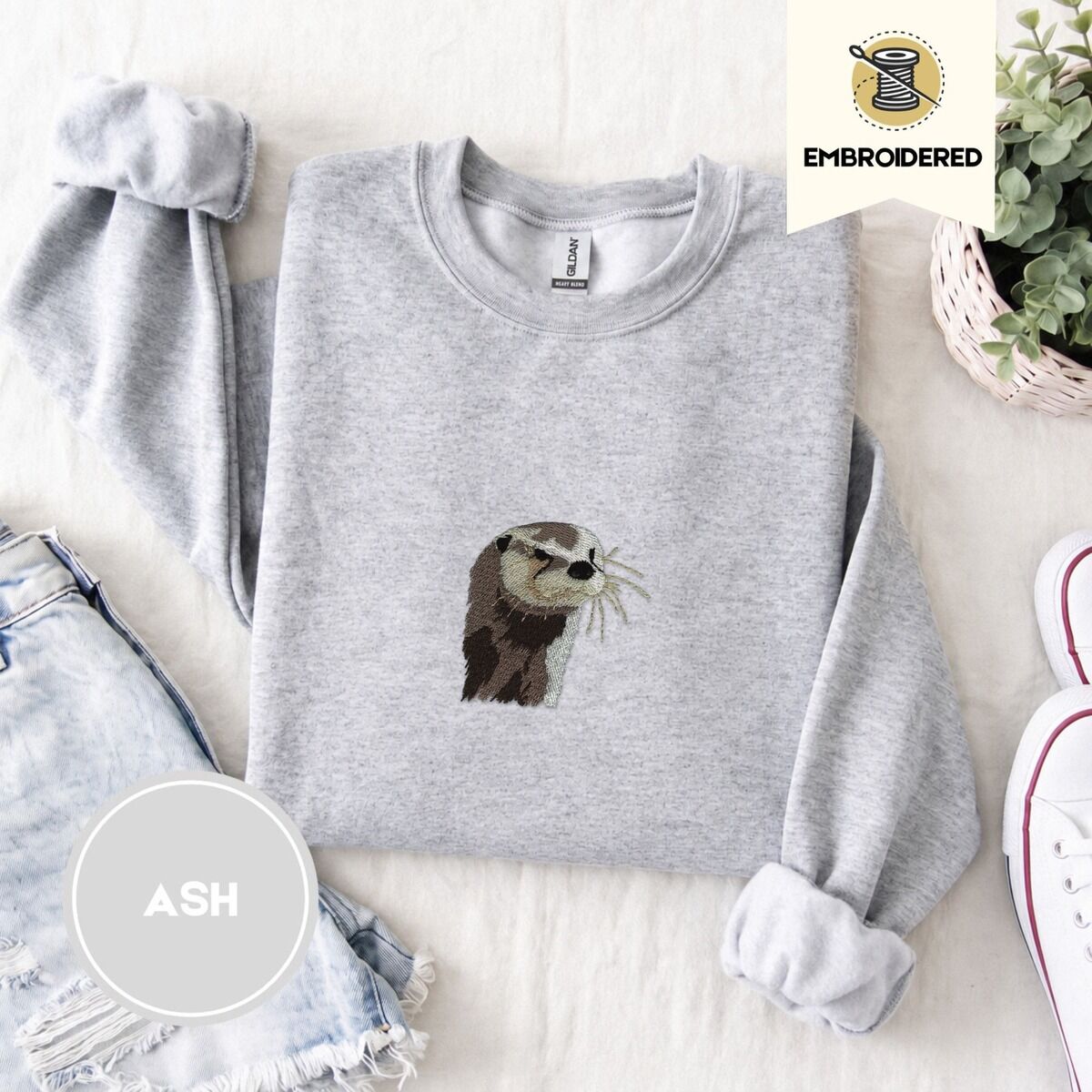 Otter Sweatshirt – Ash Ash crewneck sweatshirt folded with denim shorts, white sneakers and a potted plant, embroidered with a river otter head in brown and cream thread with long golden whiskers