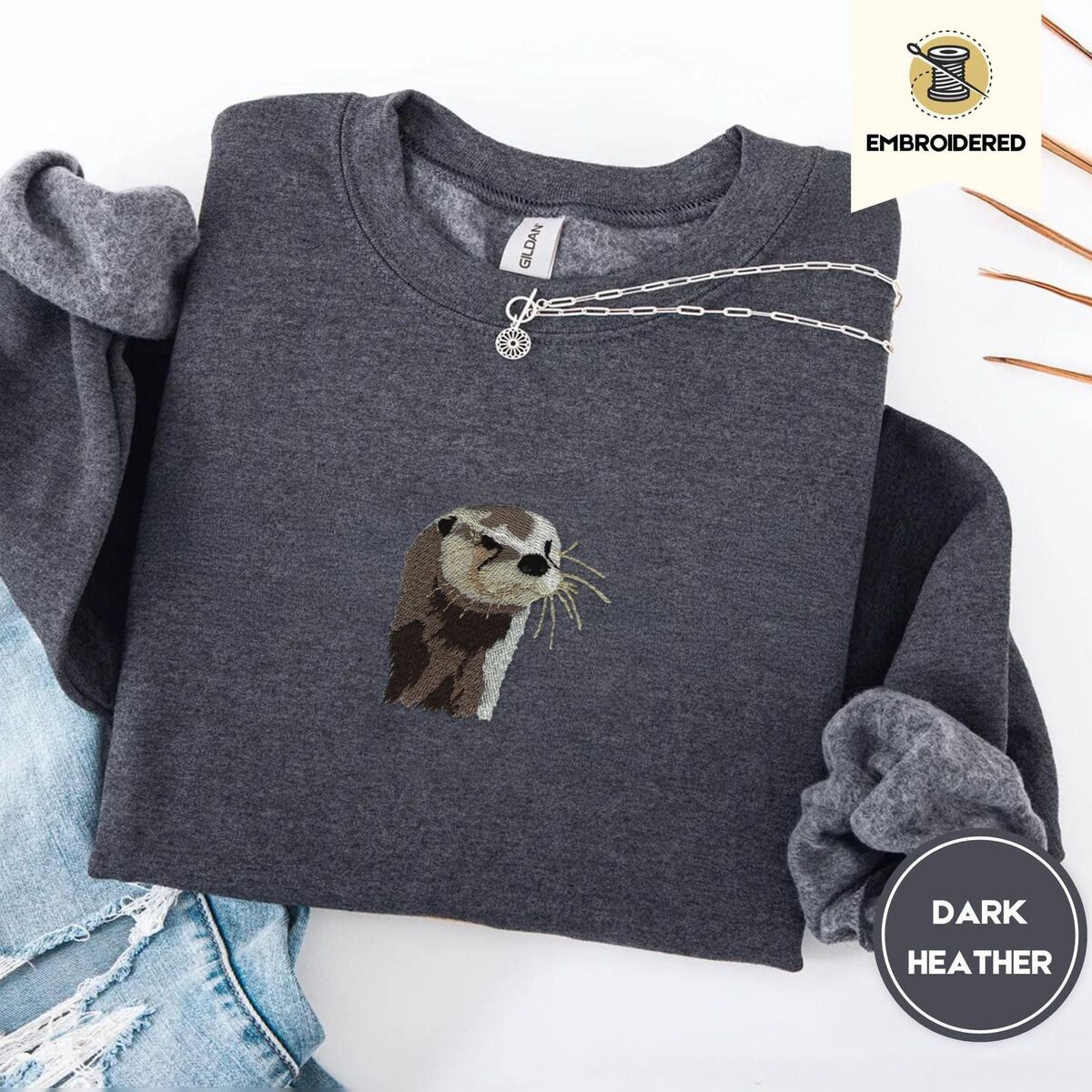 Otter Sweatshirt – Dark Heather Dark Heather crewneck sweatshirt folded with ripped jeans and a silver chain necklace, embroidered with a river otter head in brown and cream thread with long golden whiskers