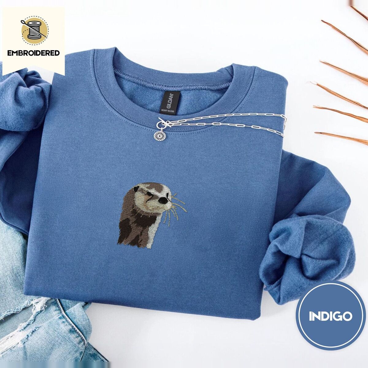 Otter Sweatshirt – Indigo Blue Indigo Blue crewneck sweatshirt folded with light jeans and dried twigs, embroidered with a river otter head in brown and cream thread with long golden whiskers