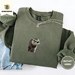 Military Green crewneck sweatshirt folded with light jeans and a silver chain necklace, embroidered with a river otter head in brown and cream thread with long golden whiskers