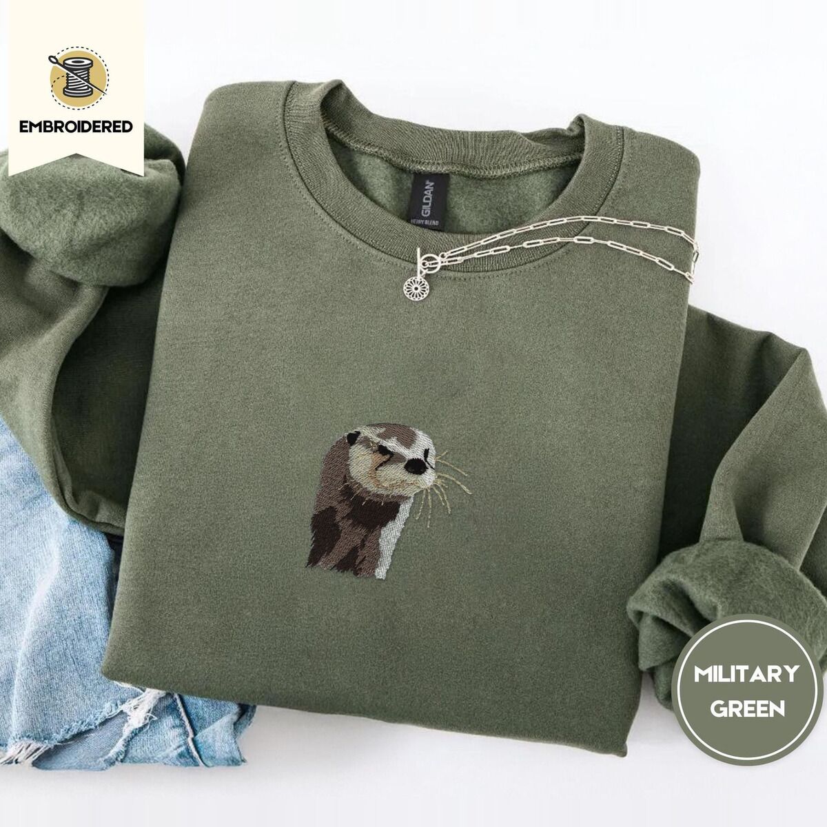 Otter Sweatshirt – Military Green Military Green crewneck sweatshirt folded with light jeans and a silver chain necklace, embroidered with a river otter head in brown and cream thread with long golden whiskers