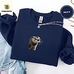 Navy crewneck sweatshirt folded with ripped jeans and a silver chain necklace, embroidered with a river otter head in brown and cream thread with long golden whiskers