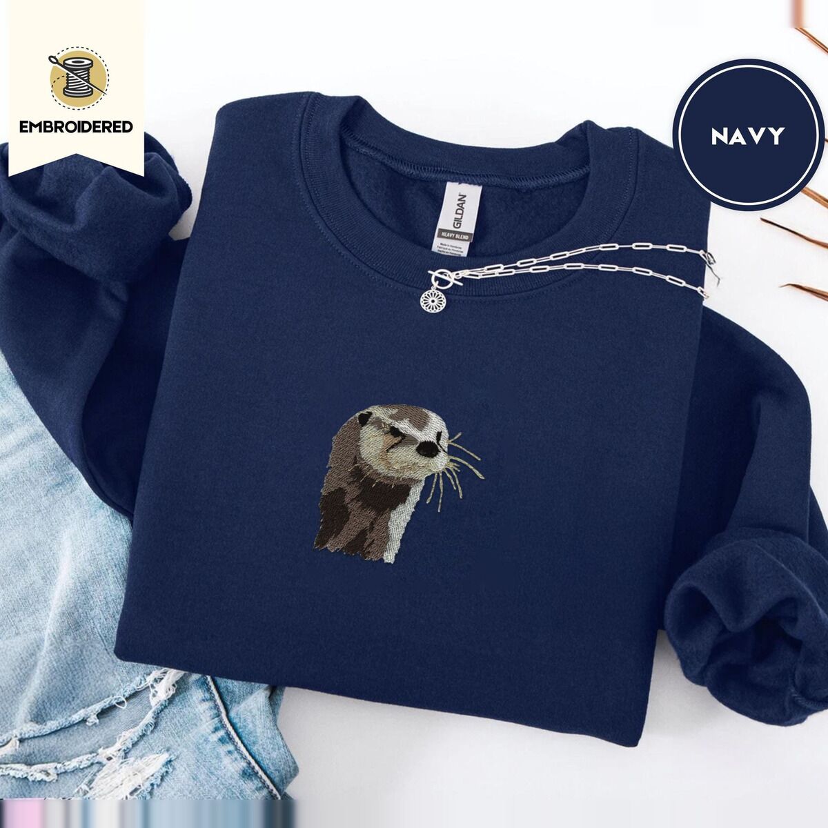 Otter Sweatshirt – Navy Navy crewneck sweatshirt folded with ripped jeans and a silver chain necklace, embroidered with a river otter head in brown and cream thread with long golden whiskers